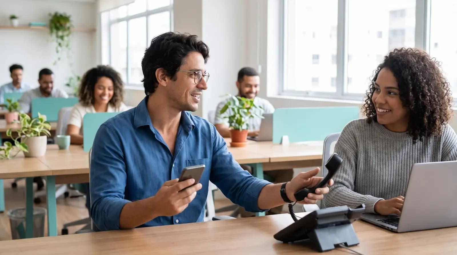 Homem de camisa azul entrega gancho de telefone para colega enquanto segura celular em escritório compartilhado.